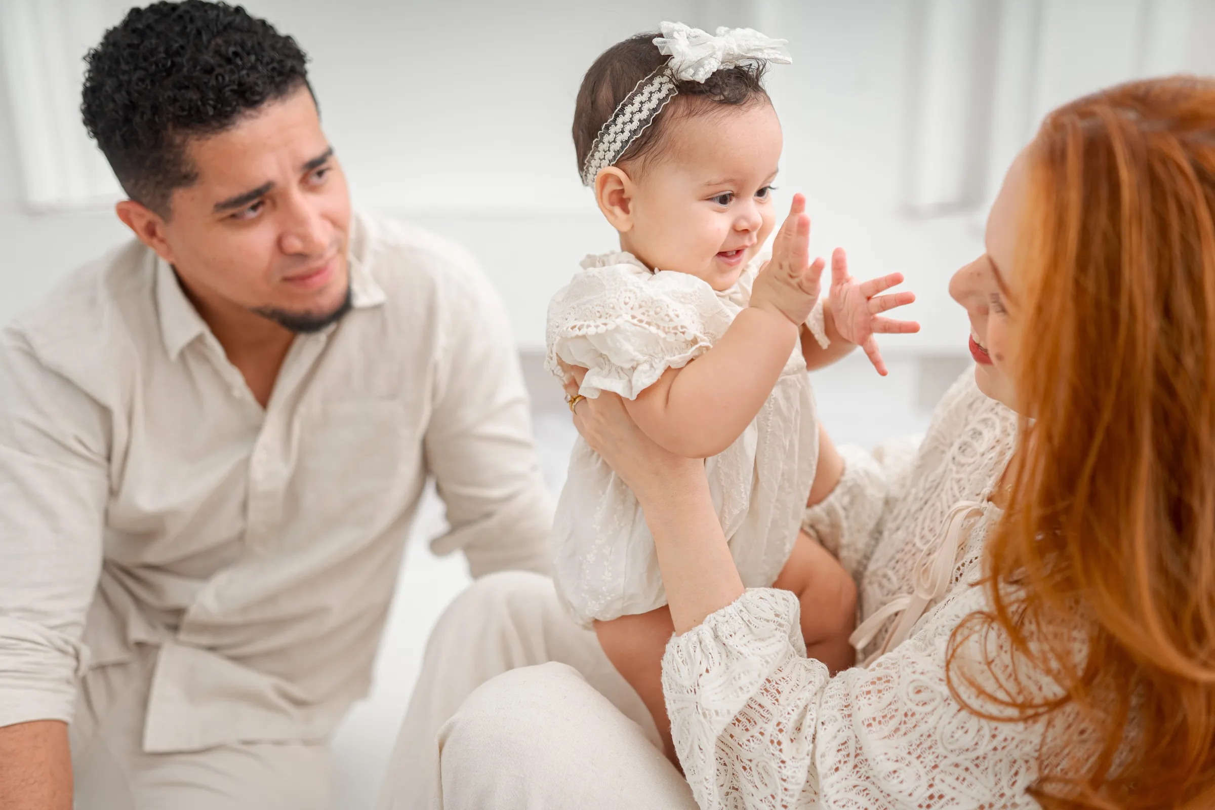 Mãe e pai rindo com a filha bebê no colo, nariz com nariz, em estúdio de luz clara